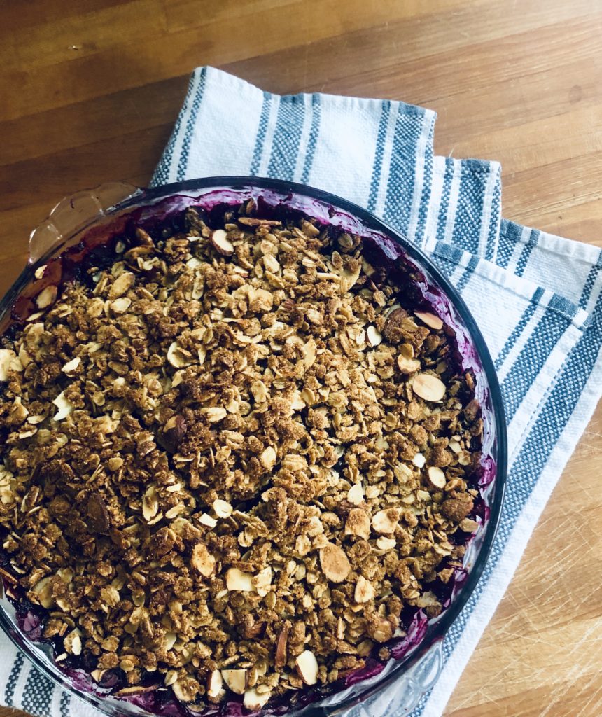 a photo of blueberry crumble in a pie plate a blue and white towel on a butcher block