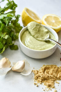 a beautiful creamy green salad dressing pictured in a small white bowl with a spoon, with green herbs, lemon, garlic cloves, and Manitoba Smooth Whole MIlled flaxseed pictured around the bowl