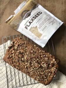 a photo of zucchini bread on a cooling rack on top of a wooden cutting board pictured next to a bag of Manitoba Milling Co. Smooth Whole Milled Flaxseed