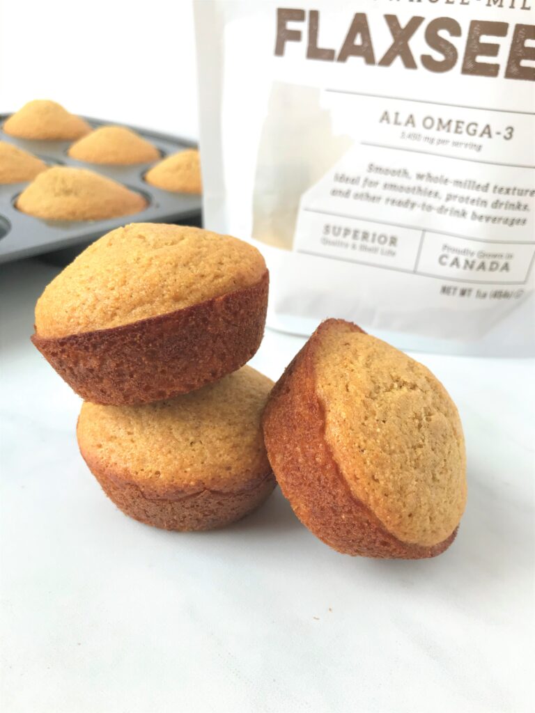 photo of three corn muffins, two are stacked on top of one another and one is balancing against the others, you can see a muffin tin in the background with more muffins as well as part of a bag of flaxseed which was used in the recipe