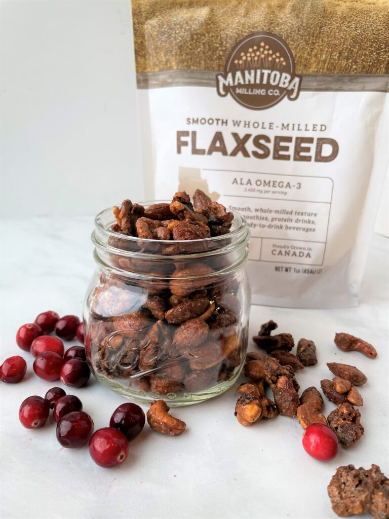 a glass mason jar filled with cinnamon sugar coated nuts, pictured on a white backdrop with red cranberries on the table and a bag of flaxseed behind